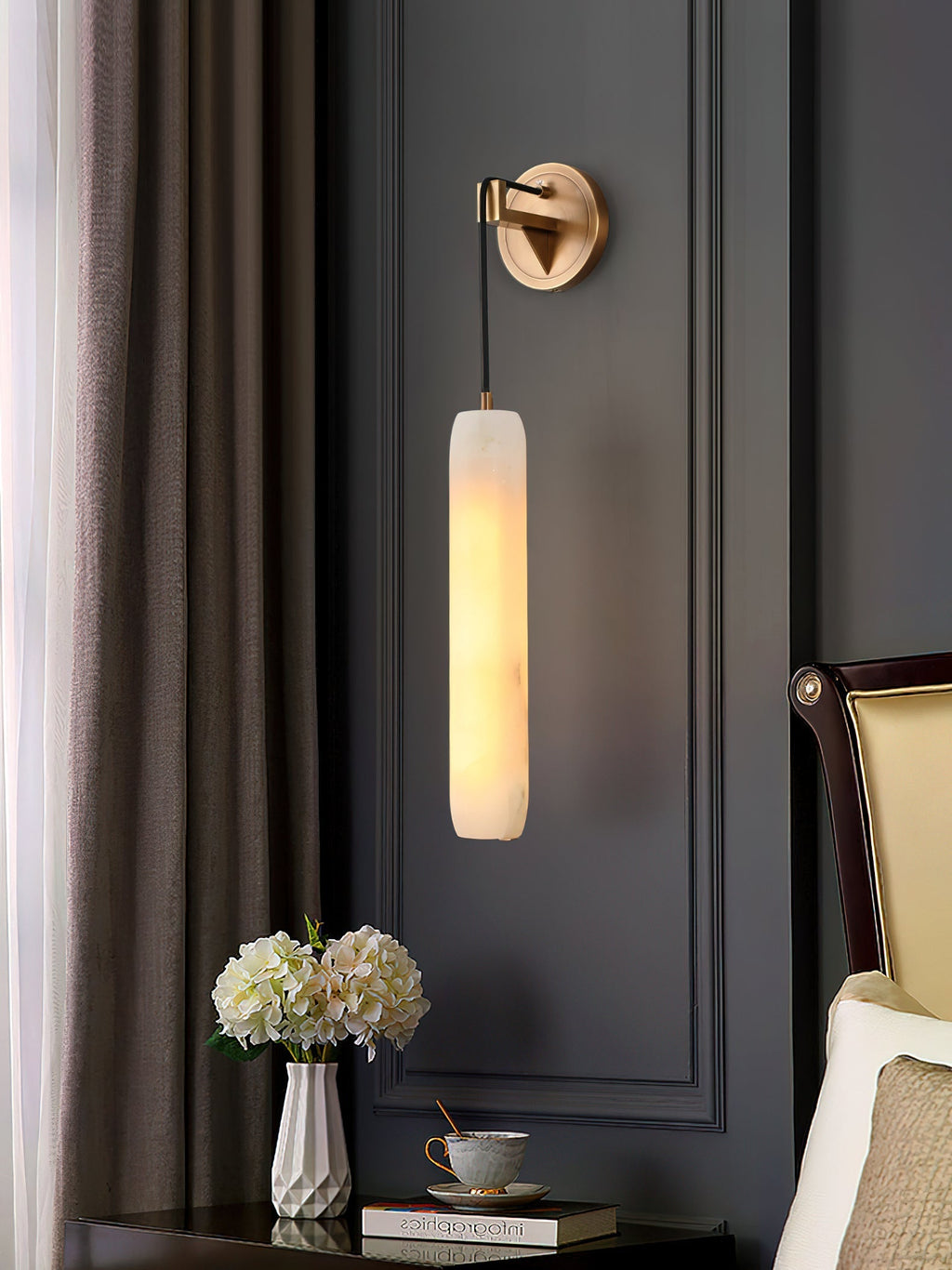 Clérachic alabaster sconce lighting a charcoal panelled bedroom wall beside a nightstand of white hydrangeas.