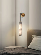 The Clérachic sconce glows against a pale plaster corner beside a dressed bed, brass plate at shoulder height.