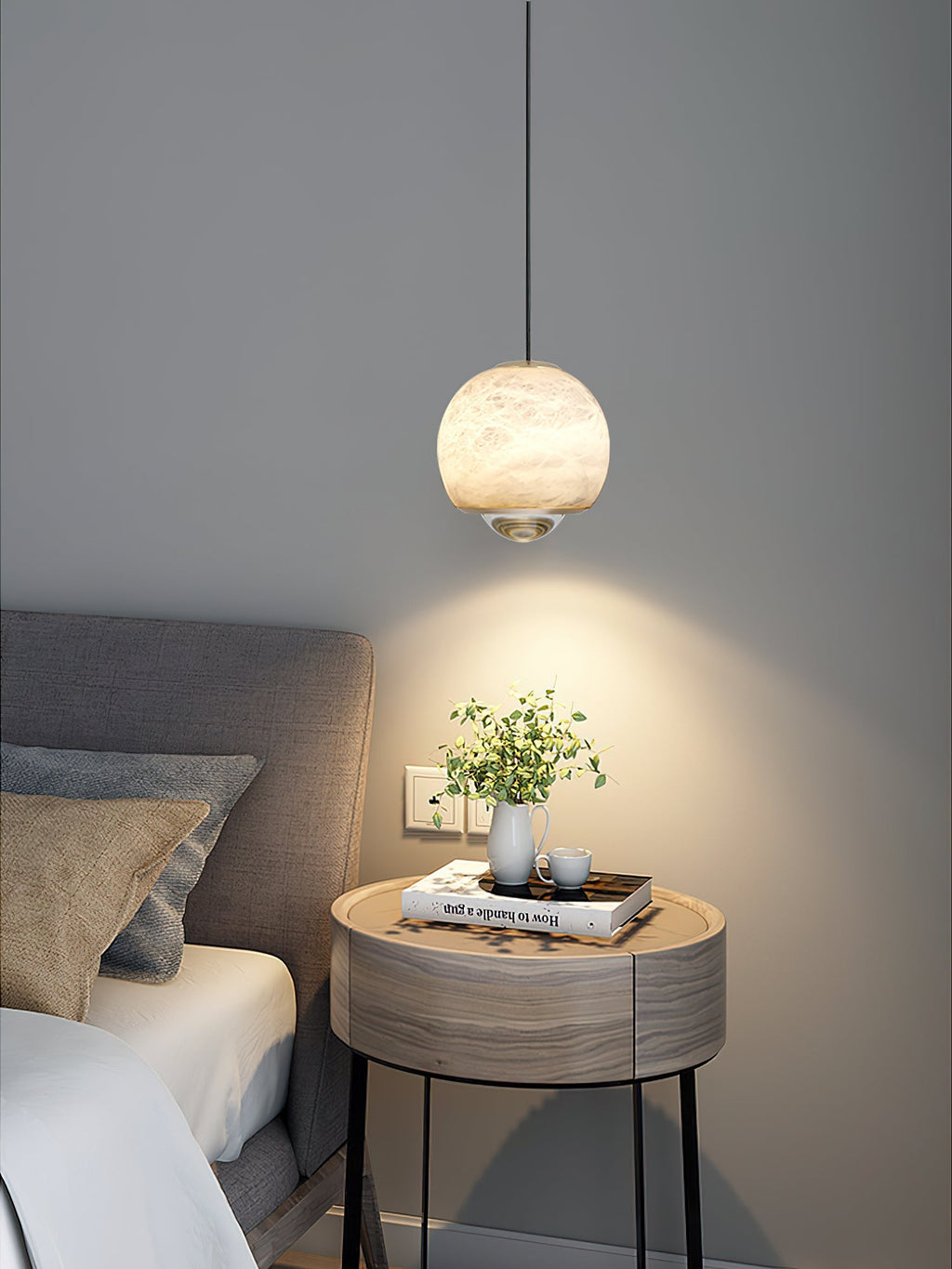 Clérachic Alabaster Globe Pendant Light lighting a round bedside table with a plant and a book, a wide warm pool spreading on the grey wall.