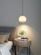 Clérachic Alabaster Globe Pendant Light lighting a round bedside table with a plant and a book, a wide warm pool spreading on the grey wall.