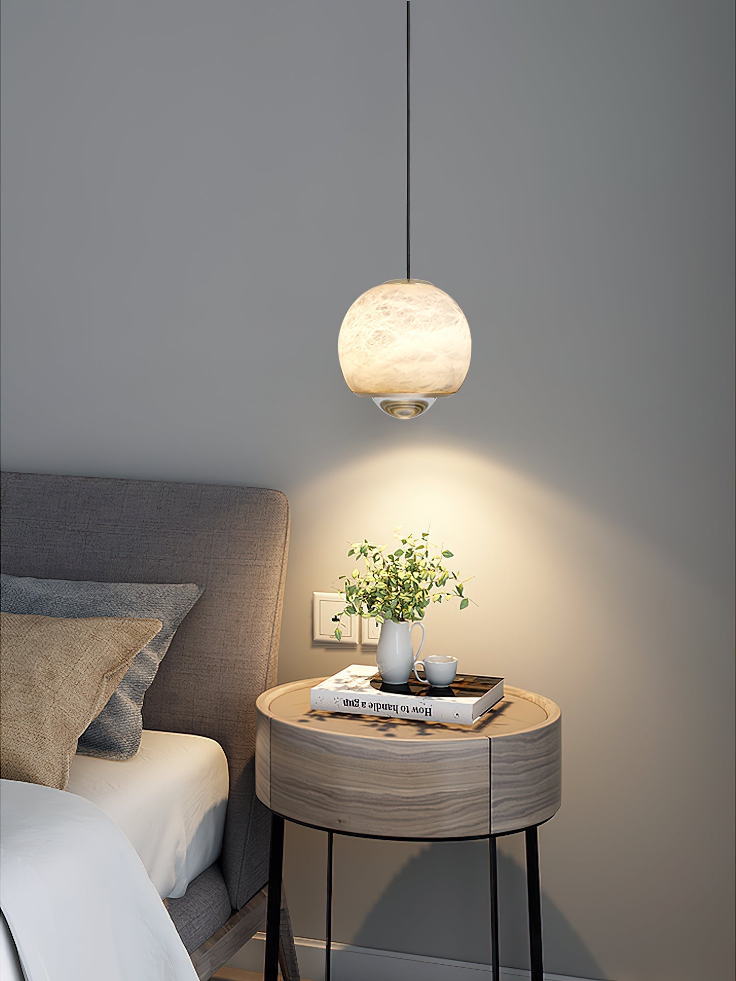 Clérachic Alabaster Globe Pendant Light lighting a round bedside table with a plant and a book, a wide warm pool spreading on the grey wall.