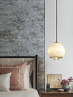 Clérachic Alabaster Globe Pendant Light lit beside a stone-clad bedroom wall, brass collar and base ring catching light above a nightstand.
