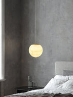 Clérachic Alabaster Globe Pendant Light on a fine cable against a raw concrete wall, glowing beside a bed and a water carafe.