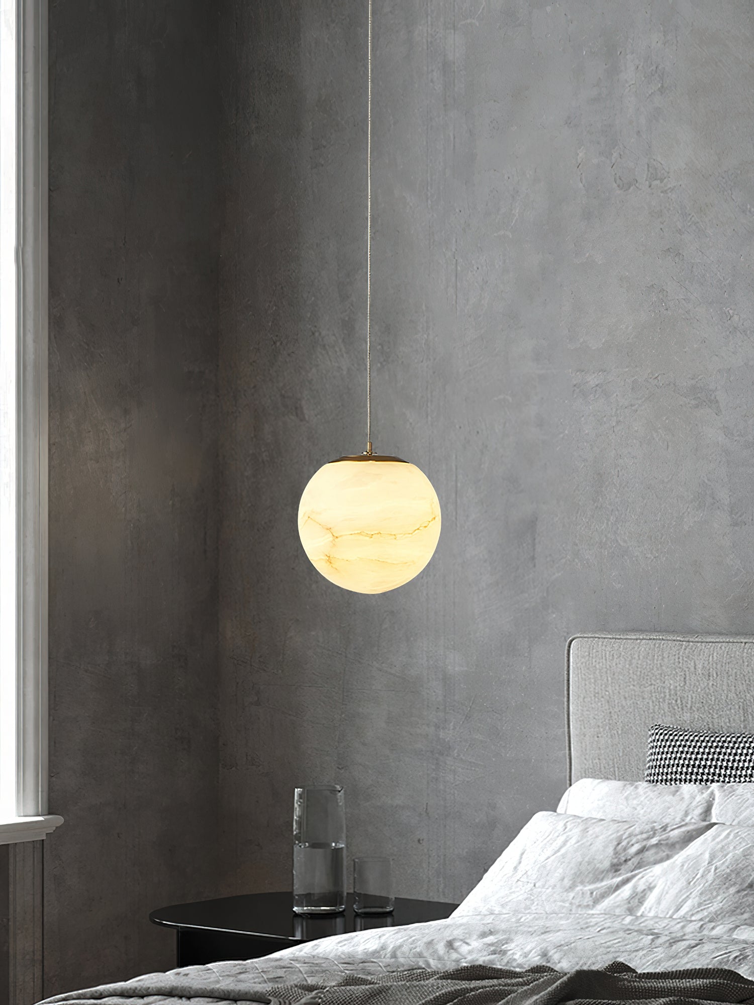 Clérachic Alabaster Globe Pendant Light on a fine cable against a raw concrete wall, glowing beside a bed and a water carafe.