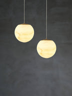 A pair of Clérachic Alabaster Globe Pendant Light globes on steel cables, lit side by side against a dark plaster backdrop.
