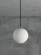 Clérachic Alabaster Globe Pendant Light on a black cord against a bare grey plaster wall, the stone reading milky and unlit.