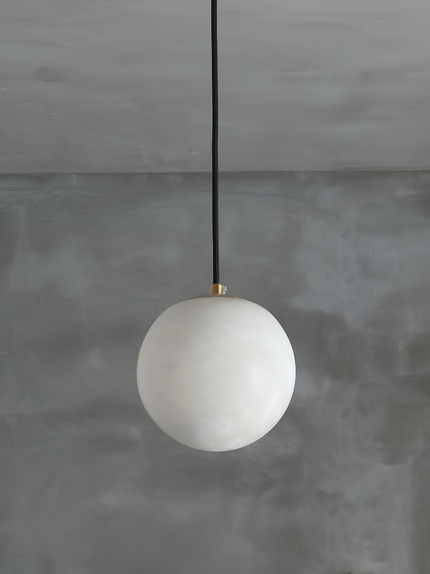 Clérachic Alabaster Globe Pendant Light on a black cord against a bare grey plaster wall, the stone reading milky and unlit.