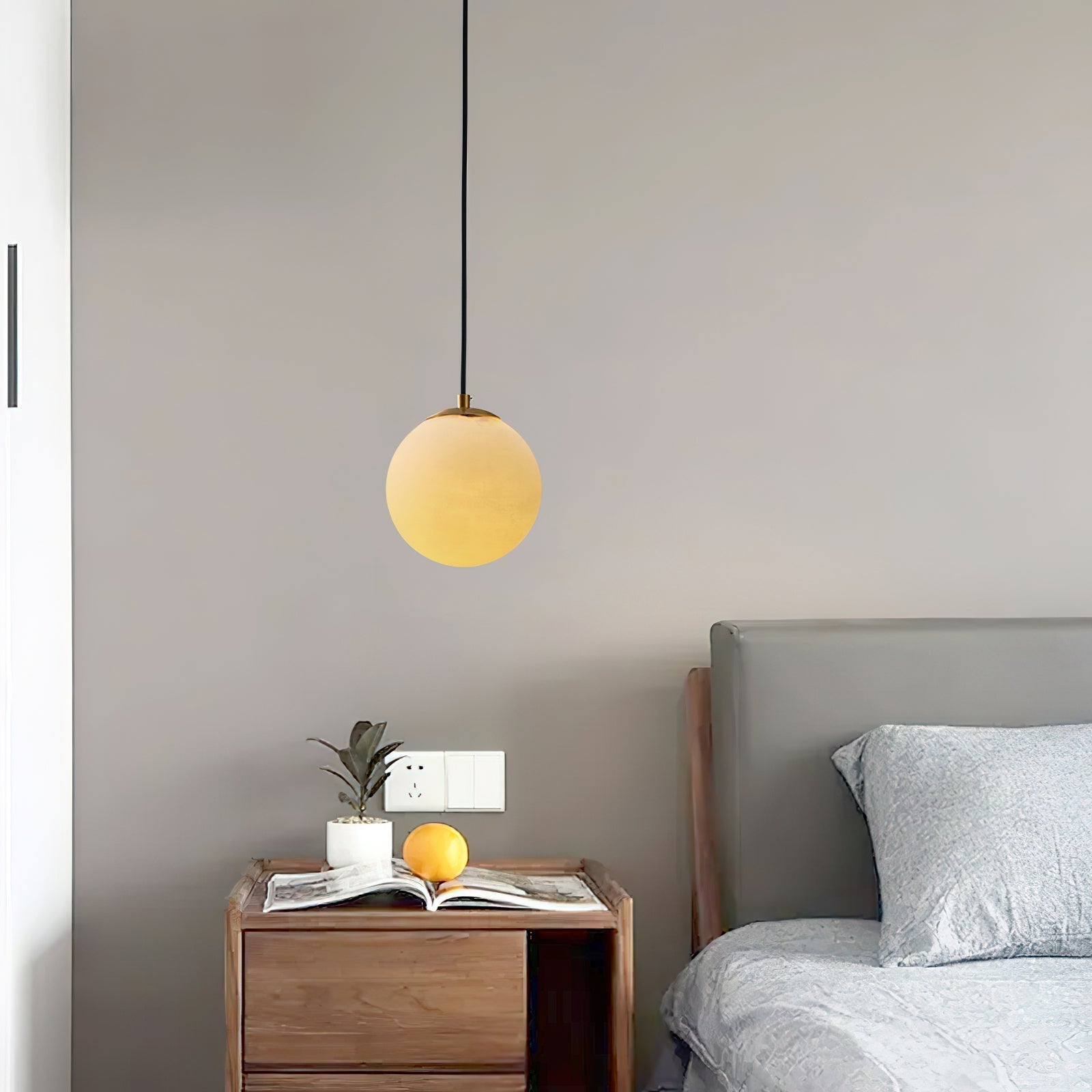 Clérachic Alabaster Globe Pendant Light glowing amber above a wooden nightstand set with a plant and an open book.