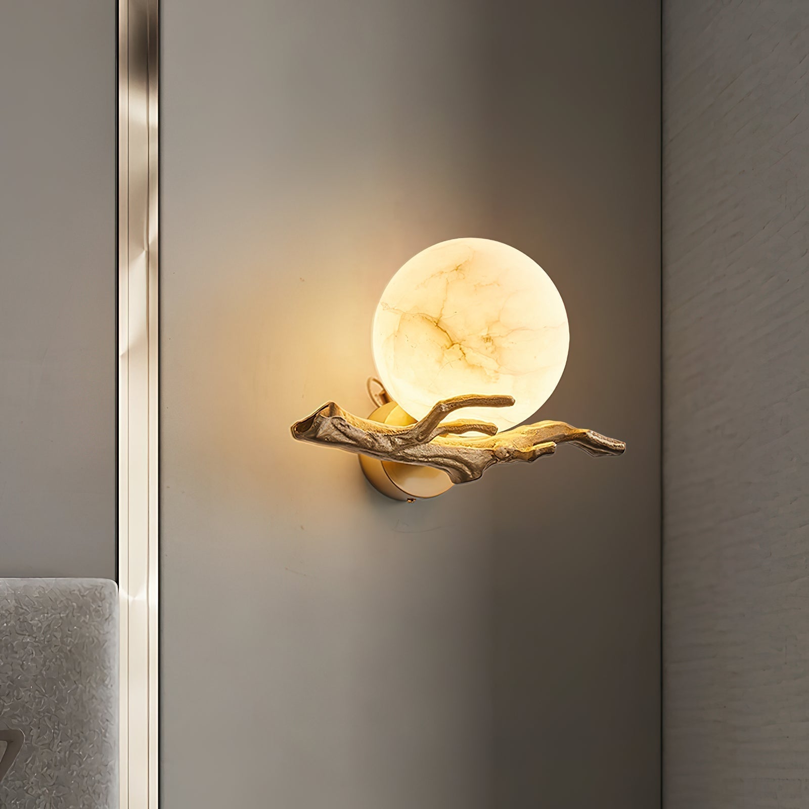 The Clérachic Alabaster Globe Wall Sconce lit on a grey wall beside a brushed metal reveal, its branch arm cast in fine relief.