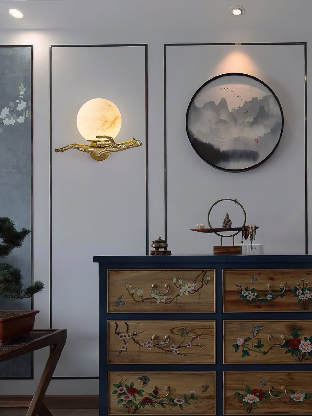 The Clérachic Alabaster Globe Wall Sconce on a white panelled wall above a painted chest, its globe echoing a round ink painting.