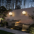 Two Clérachic Alabaster Globe Wall Sconces lit along a stone wall above a tiered water feature at dusk, bamboo rising behind.