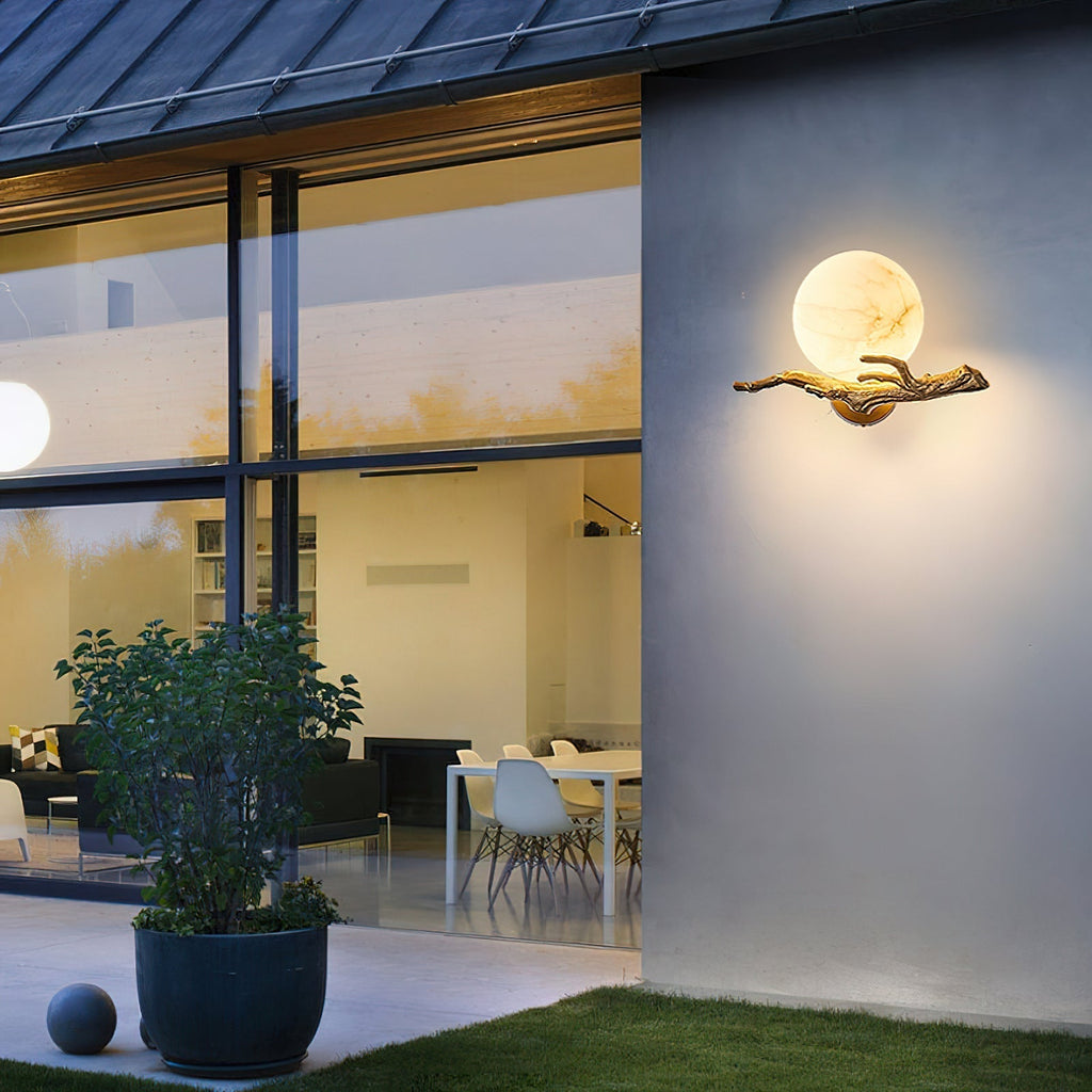 Clérachic Alabaster Globe Wall Sconce lit on a rendered wall at dusk, a glazed room and lawn beside it in the same frame.