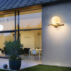 Clérachic Alabaster Globe Wall Sconce lit on a rendered wall at dusk, a glazed room and lawn beside it in the same frame.