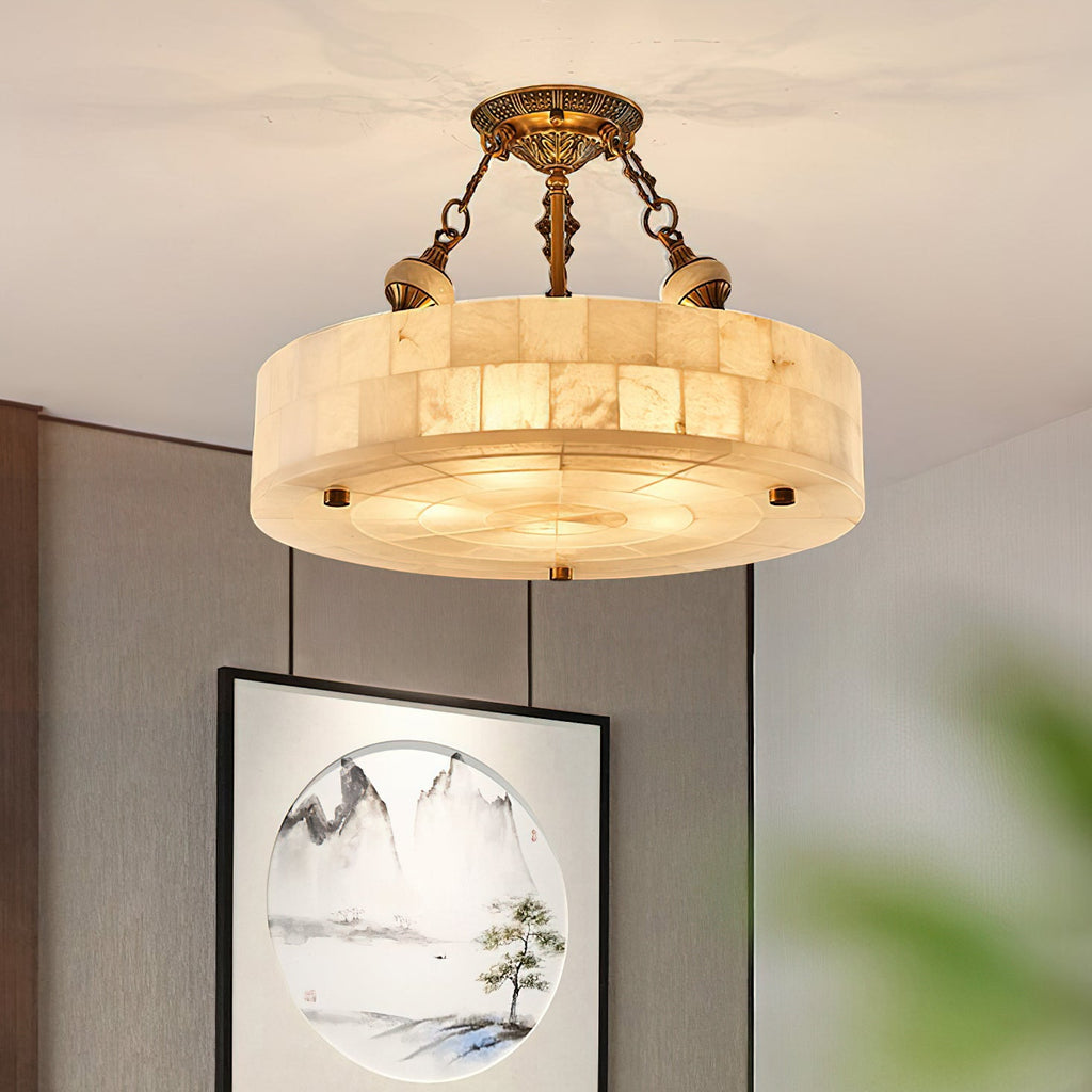 Lit alabaster drum Clérachic ceiling light with antique brass chains, beside a framed mountain landscape print on a grey wall.