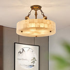 Alabaster drum Clérachic ceiling light lit in warm tones, hanging in front of a grey wall with a framed landscape picture.