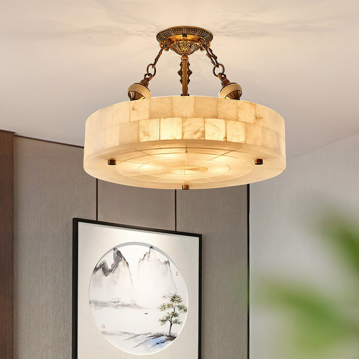 Alabaster drum Clérachic ceiling light lit in warm tones, hanging in front of a grey wall with a framed landscape picture.
