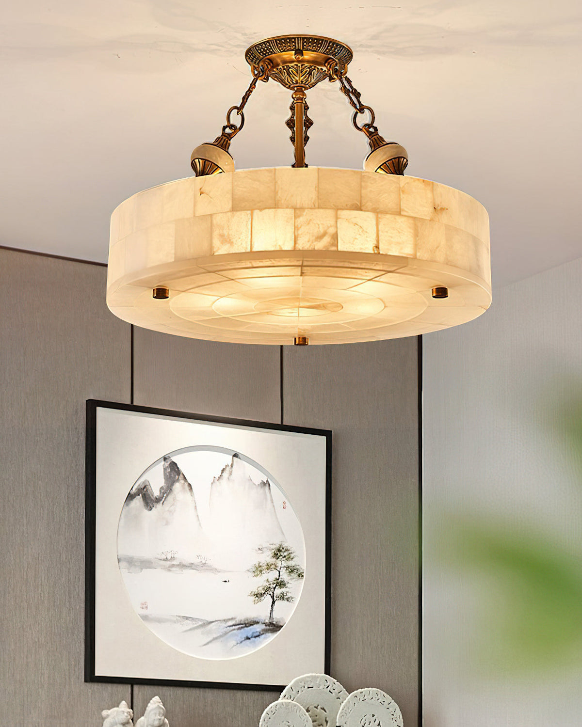 This Clérachic ceiling light hangs in front of a grey wall, its alabaster blocks lit from inside, with a framed landscape print below.