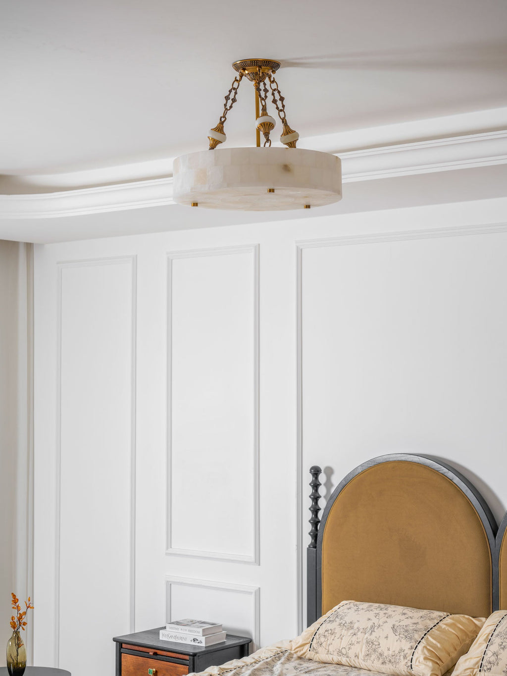 Clérachic alabaster ceiling light hanging below a cornice in a bedroom, with a black-framed headboard and a bedside table to one side.