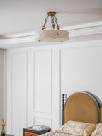 Clérachic alabaster ceiling light hanging below a cornice in a bedroom, with a black-framed headboard and a bedside table to one side.