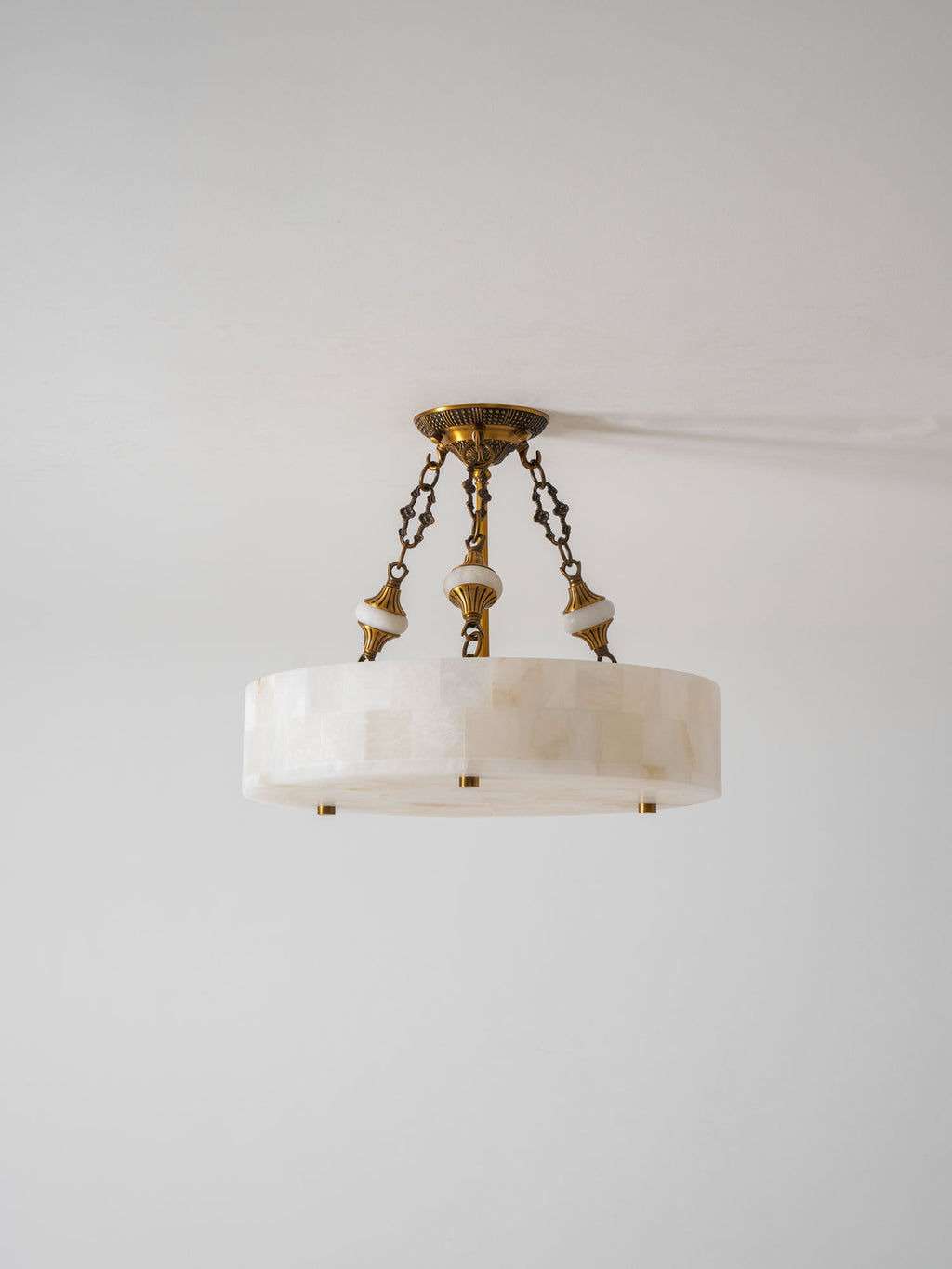 Slim alabaster drum Clérachic ceiling light with three chains and brass studs, photographed against a pale grey wall.