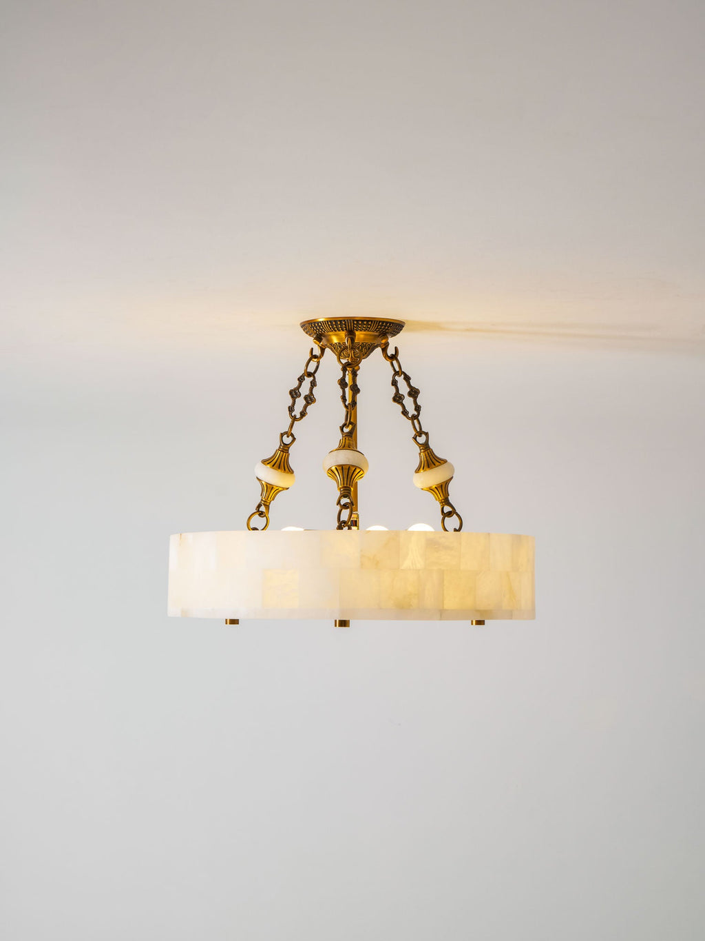 Lit Clérachic alabaster ceiling light with three brass chains, a warm glow at the ceiling line and a pale grey wall behind.