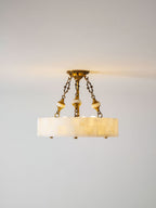 Lit Clérachic alabaster ceiling light with three brass chains, a warm glow at the ceiling line and a pale grey wall behind.