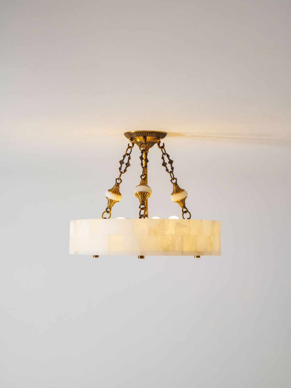 Lit Clérachic alabaster ceiling light with three brass chains, a warm glow at the ceiling line and a pale grey wall behind.