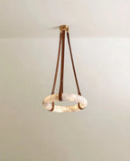 Clérachic alabaster ring pendant on three tan leather straps and a gold canopy, glowing softly against a pale wall.