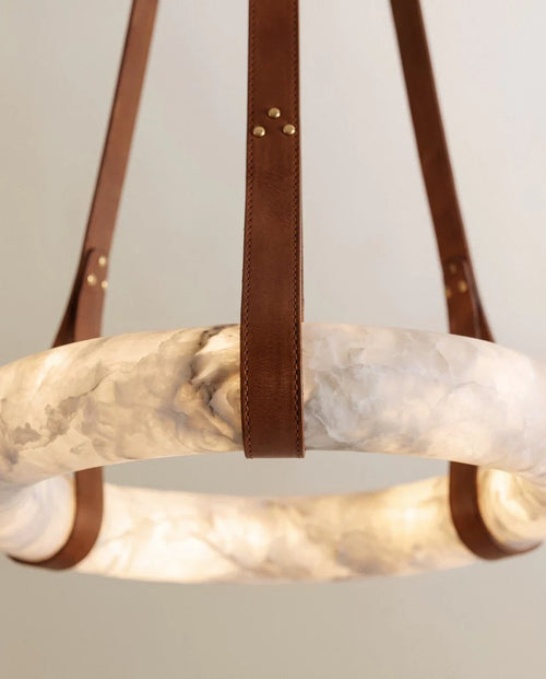 Close view of the Clérachic alabaster ring, its cloudy veining lit from within, held by a stitched tan leather strap with gold rivets.