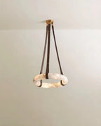 Clérachic alabaster ring pendant hung from three dark brown leather straps and a gold canopy, lit against a pale wall.