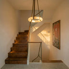 Clérachic alabaster ring pendant on leather straps hanging in a stairwell above a walnut staircase, with warm evening light.