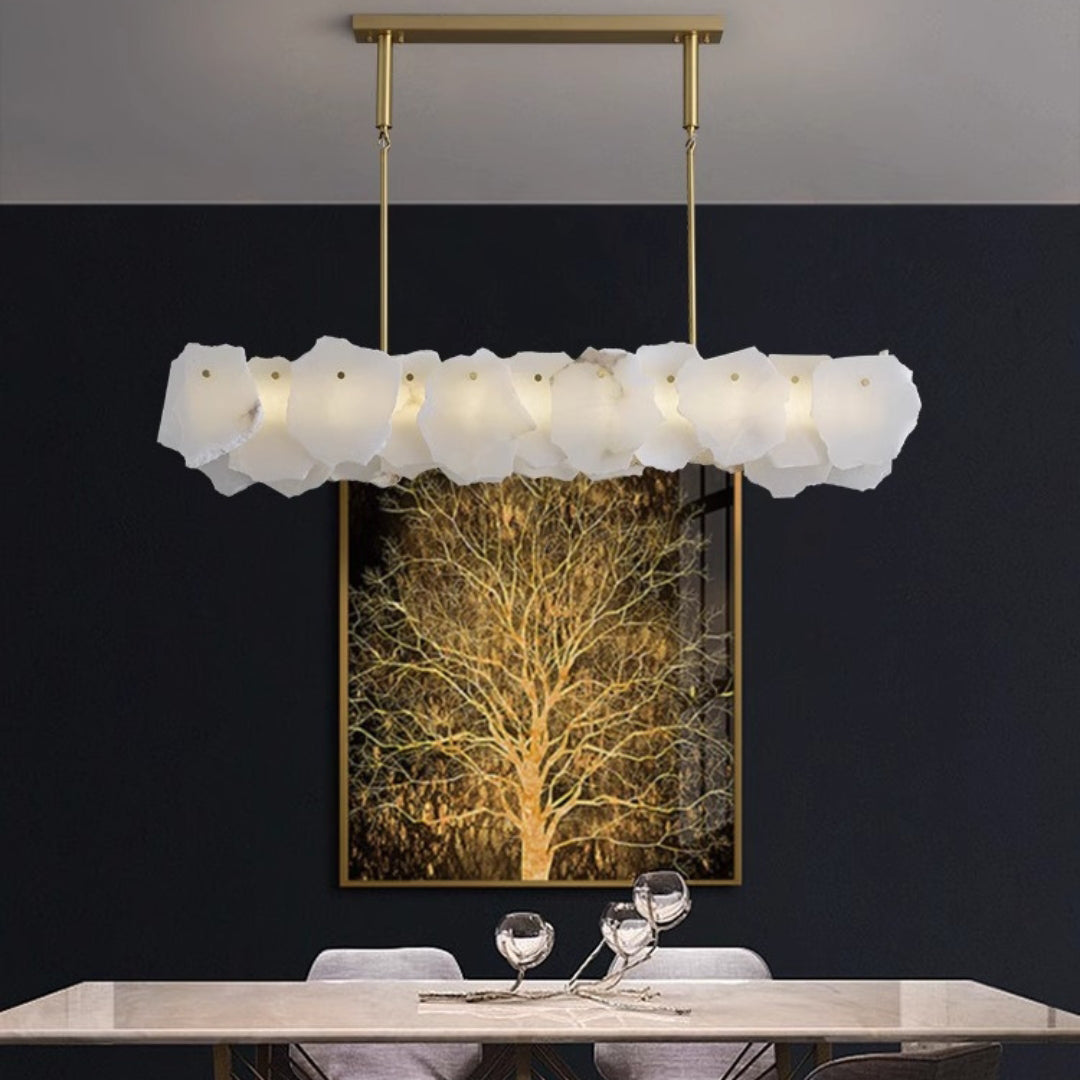 Clérachic alabaster linear chandelier lit against a dark wall hung with a gilded tree canvas above a stone table
