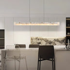The Clérachic linear light above a kitchen island, warm glow against a veined marble splashback and bar stools.