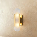 Clérachic wall light with a pill-shaped alabaster shade held by gold metal bars on a gold wall plate.