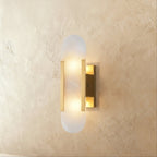 Clérachic wall light with a pill-shaped alabaster shade held by gold metal bars on a gold wall plate.