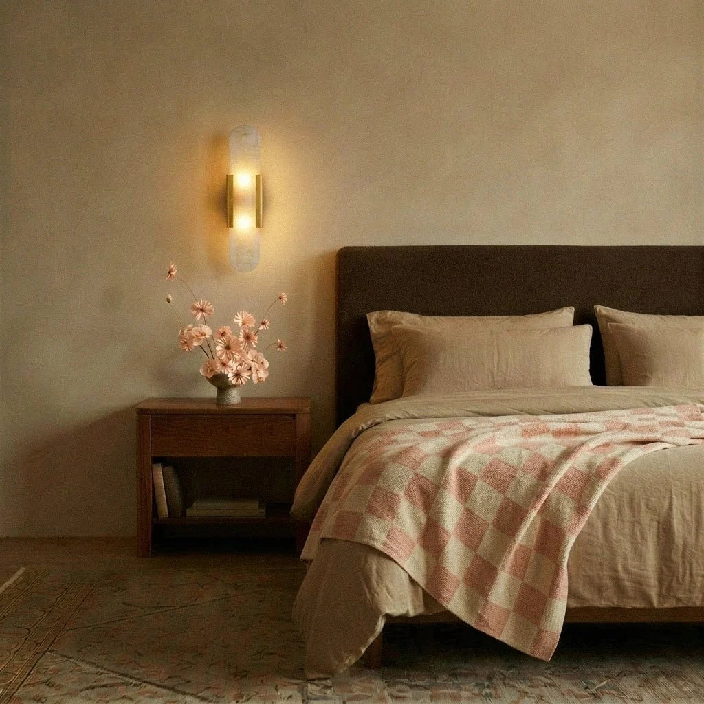 Clérachic gold wall light with an alabaster shade glowing above a walnut bedside table in a calm bedroom.