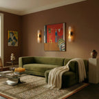 Two Clérachic alabaster wall lights lit either side of a painting above a green velvet sofa in a living room.