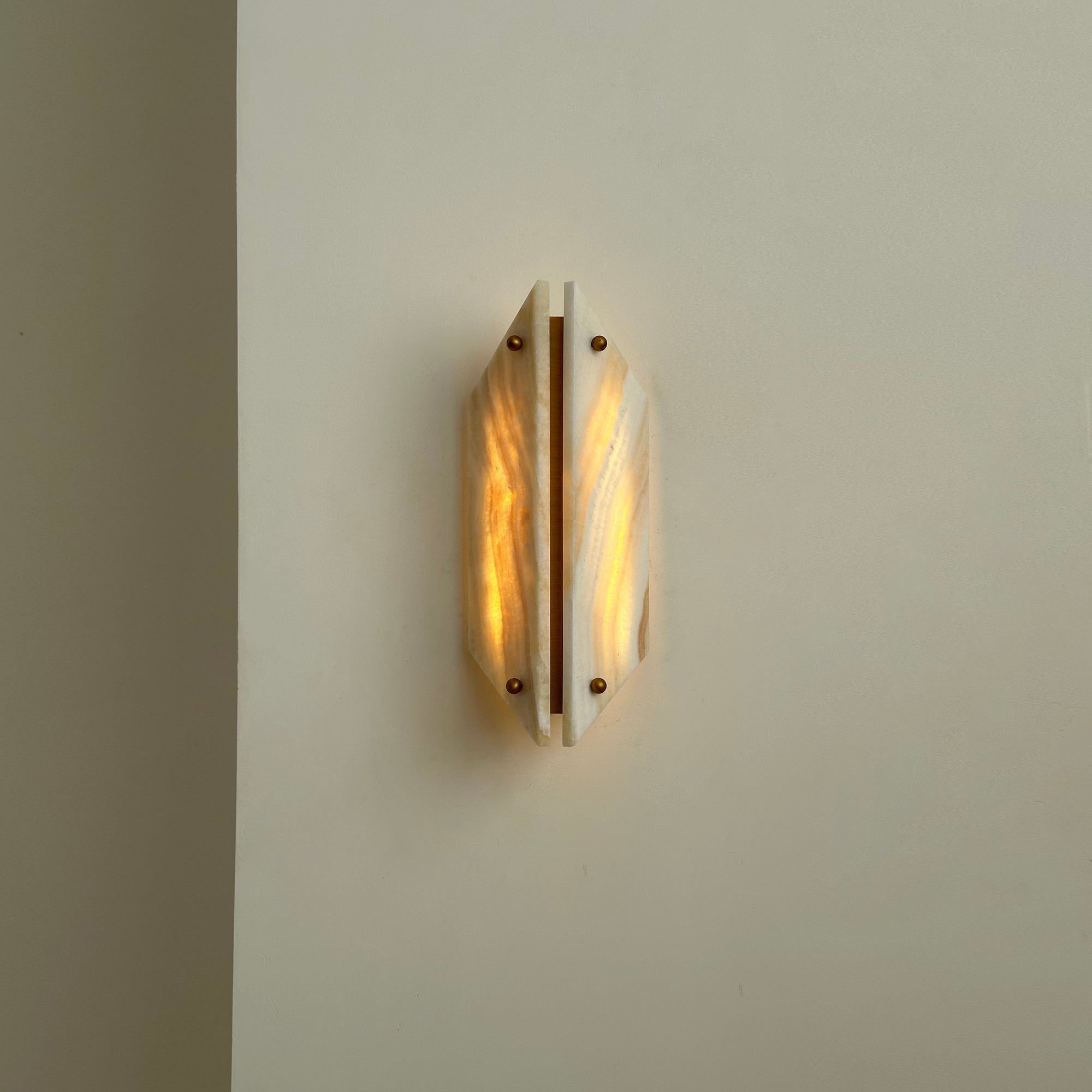Clérachic alabaster sconce lit on a cream wall, both stone panels glowing warm with a bright brass strip between.