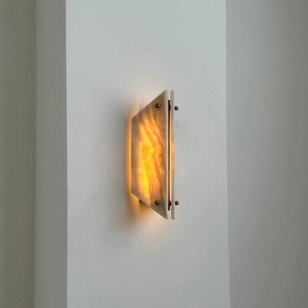 Clérachic stone sconce lit on a white hallway wall, a narrow blade of light escaping the gap between the panels.