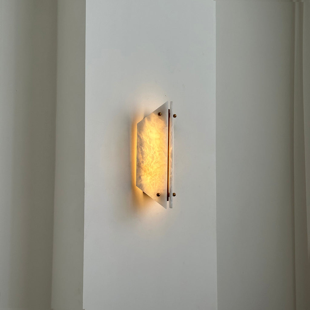 The Clérachic alabaster sconce lit in a tall hallway, a small pool of warm light on the plaster beside it.