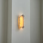 The Clérachic alabaster sconce lit in a tall hallway, a small pool of warm light on the plaster beside it.