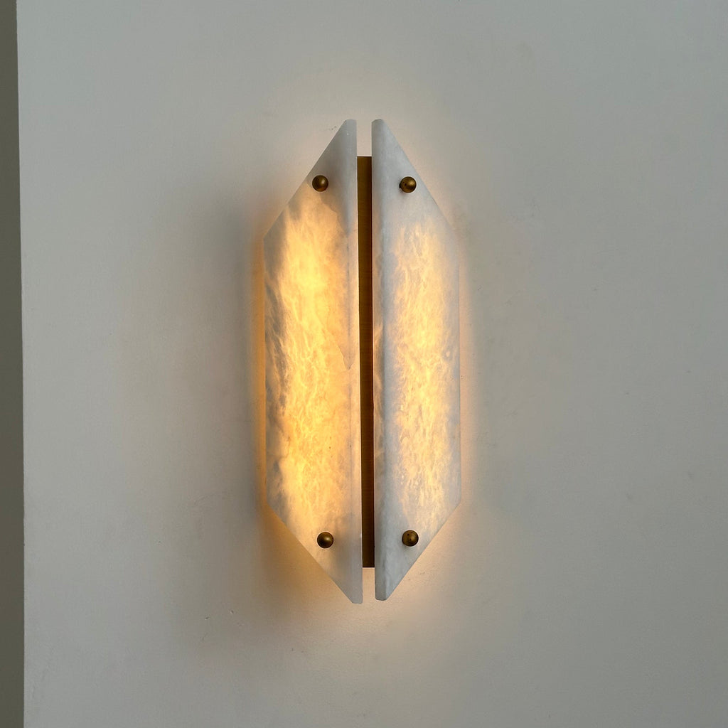Clérachic sconce lit beside a wall corner, warm light rising from behind the two angled alabaster panels.