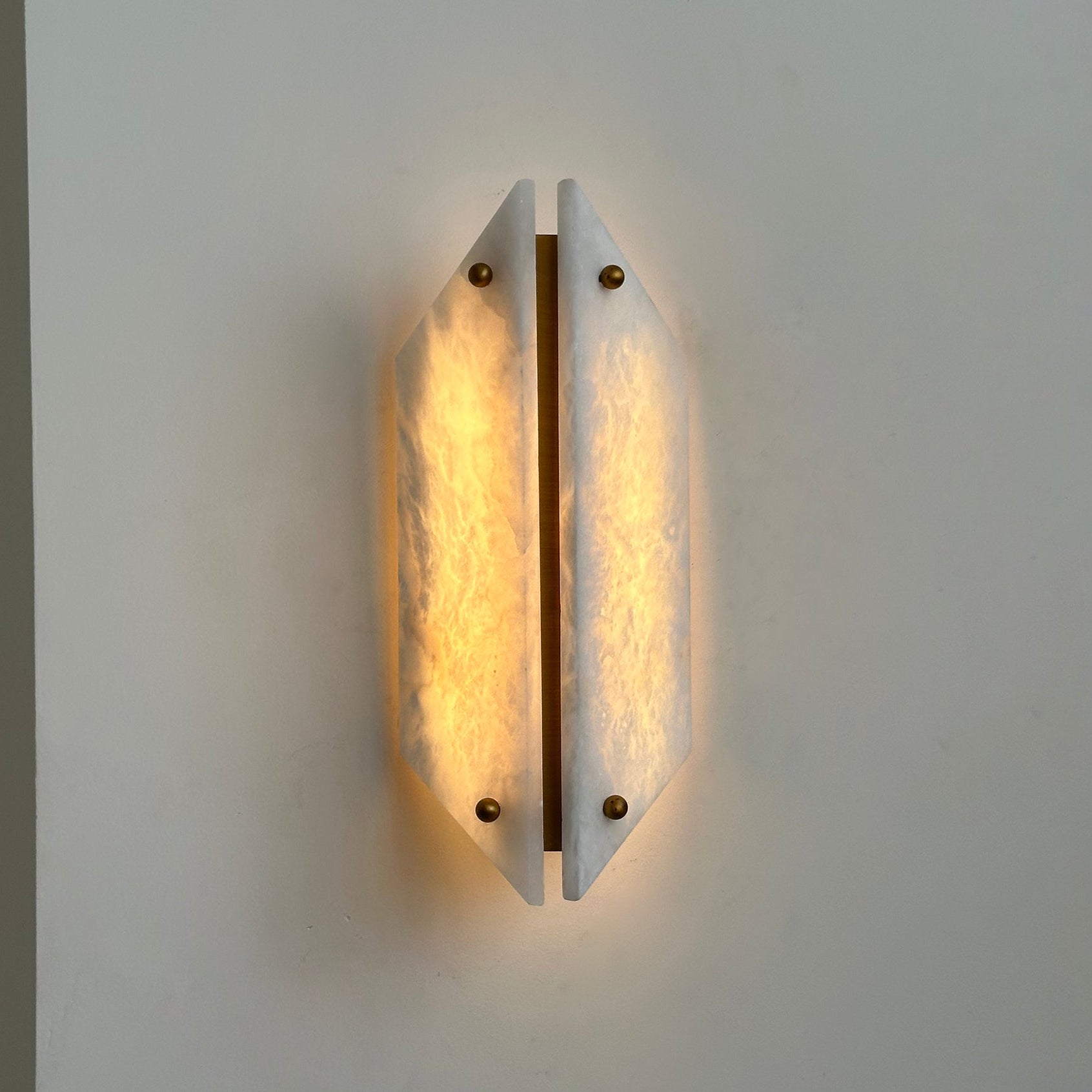 Clérachic sconce lit beside a wall corner, warm light rising from behind the two angled alabaster panels.