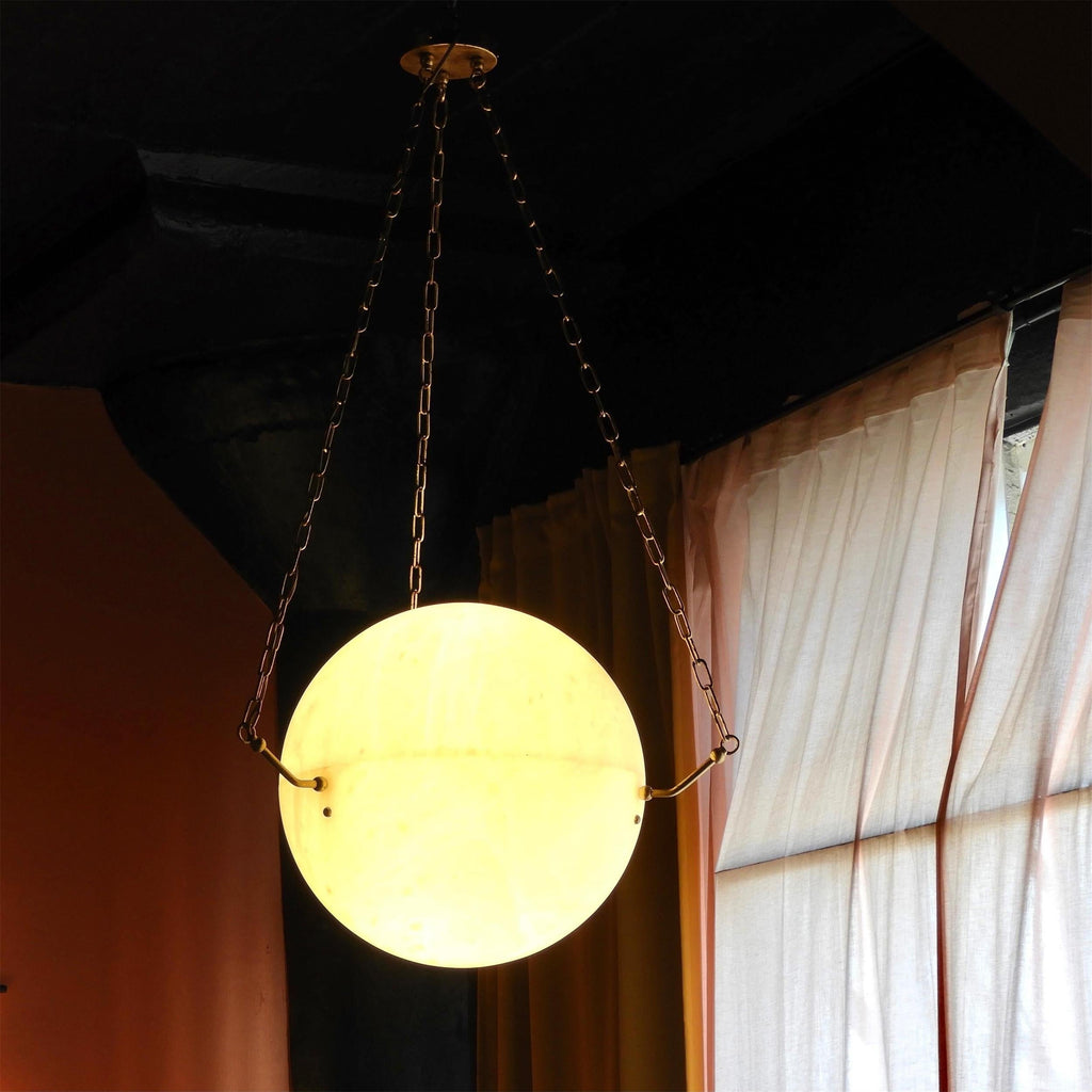 The Clérachic pendant lit in a dark room, the alabaster glowing amber beside a curtained window.