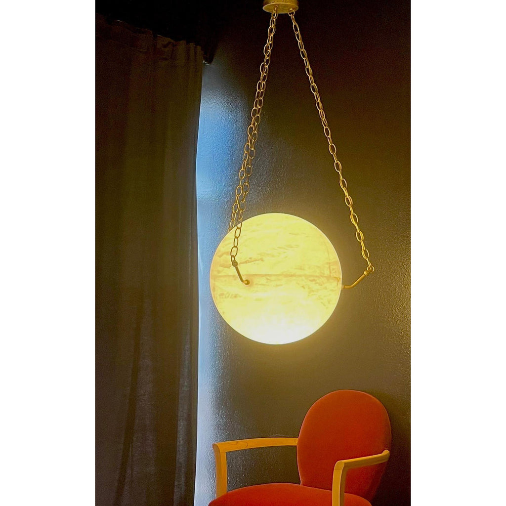Clérachic pendant glowing warm amber against a dark curtain, an orange armchair below in shadow.