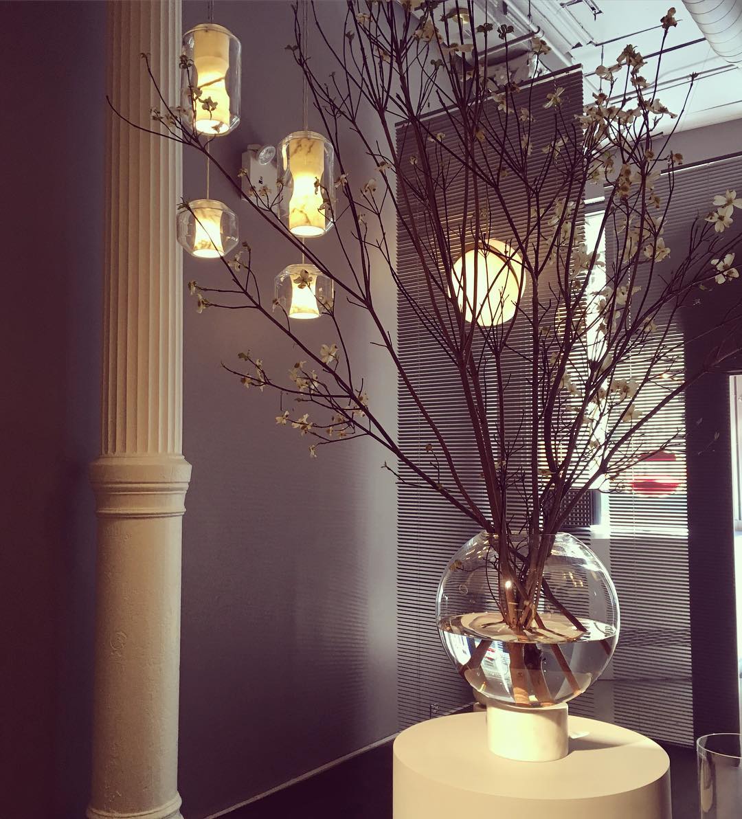 Four Clérachic Alabaster Pendant Lights lit beside a fluted column, seen through the blossom branches of a tall vase.