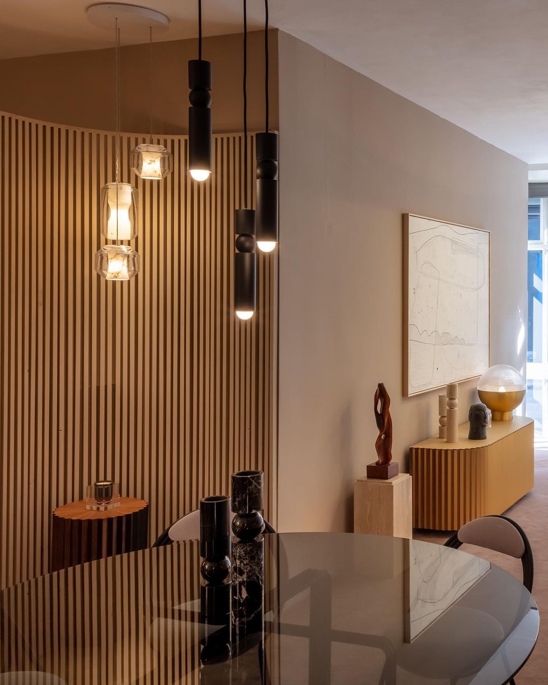 Three Clérachic Alabaster Pendant Lights lit over a round glass dining table against a curved slatted oak wall.