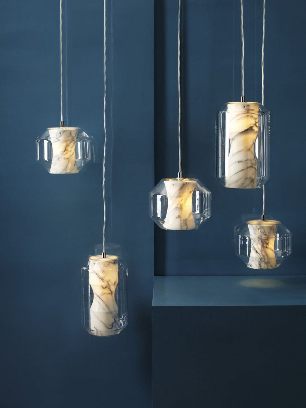 Five Clérachic Alabaster Pendant Lights lit against a deep blue wall, the grey veining in each stone core reading clearly.