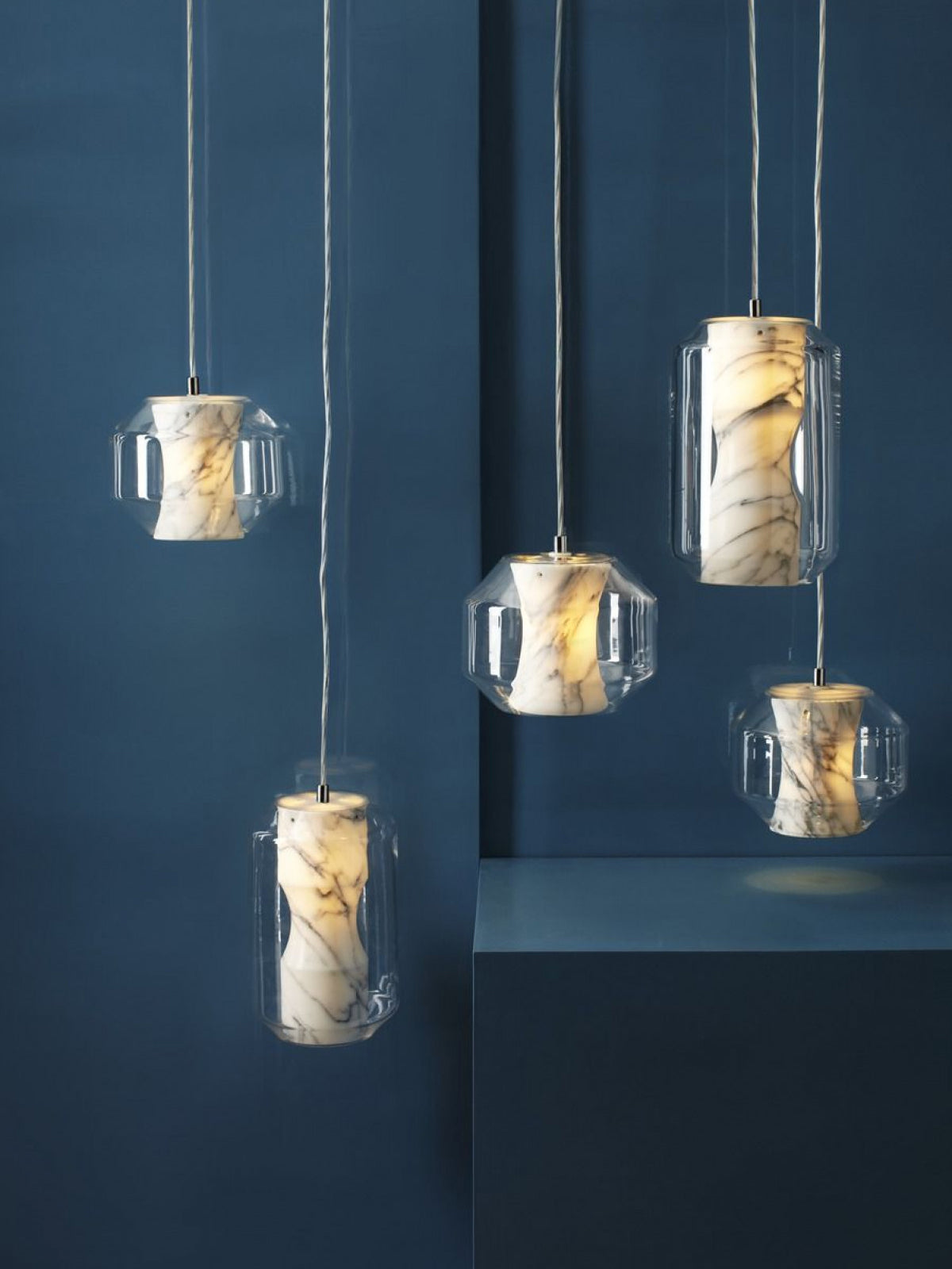 Five Clérachic Alabaster Pendant Lights lit against a deep blue wall, the grey veining in each stone core reading clearly.