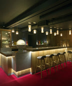 A long run of Clérachic Alabaster Pendant Lights lit above a timber cocktail bar and its row of pale oak stools.